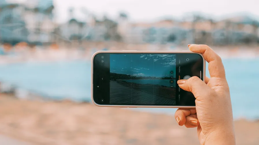 En hånd holder en smartphone på tværs og fotograferer en strand med hav i baggrunden