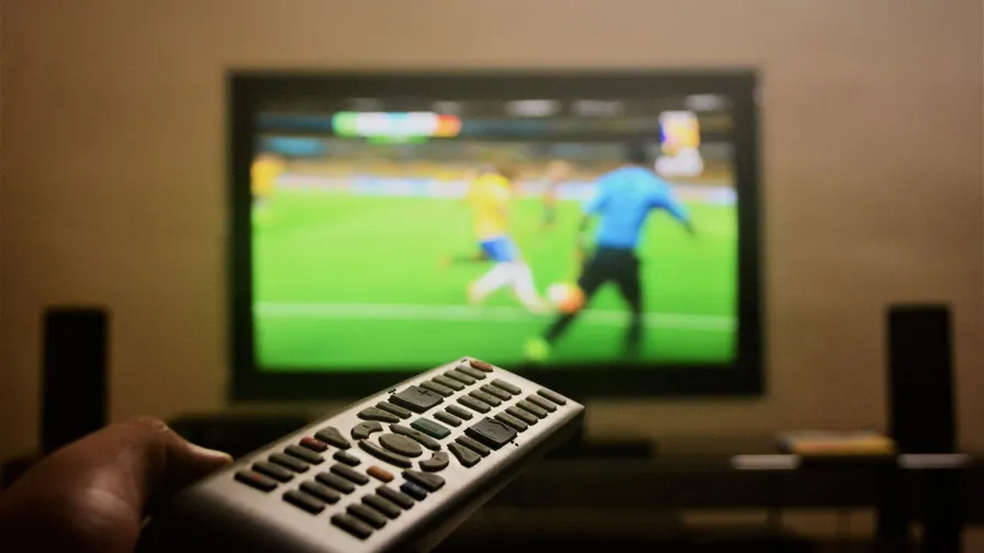 En hånd holder en fjernbetjening foran et tv, der viser en fodboldkamp